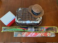 Picnic basket, small cooler, sun hat, and two kites packed in plastic sleeves shown on wood surface.