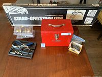 Overview of the lot items: Ladder Pro stand-off/stabilizer box, red metal toolbox, door knobs in packaging, hand tools, and nails on wood table.