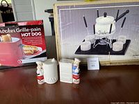 Overall view of all items in the lot including fondue pot with forks and stand, hot dog toaster box, and salt and pepper shakers on table.
