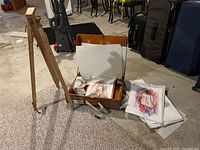 Wooden easel set up with artist's supply box open showing paintings and art materials inside. Some unfinished watercolor artworks and scattered papers visible.