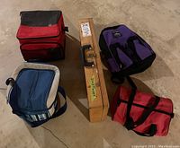 Photo showing one small purple backpack, one red and black soft cooler bag, one blue soft cooler bag with striped sides, a red duffel bag, and a rectangular wooden box with handle and latch.