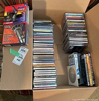 Box containing over 40 music CDs and DVDs along with several VHS tapes. Visible are multiple stacks of jewel case CDs and media in paper sleeves.
