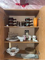 Three shelves of cabinet showing assorted dishes including brown ceramic soup pots, glass ramekins, ceramic mugs, white gravy boat, plates, serving trays, and cake stand.