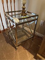 Front angle view of metal frame end table with clear glass top and bottom shelf, metal crossed side design, and caster wheels.