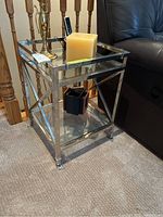 Metal frame end table with glass top and bottom shelves shown near recliner chair and staircase railing.