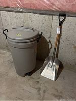 Photo of beige Rubbermaid garbage bin with lid and black wheels beside a metal shovel with wooden handle and black D-grip handle, placed on a concrete floor against a concrete wall.