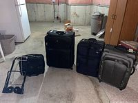 Four pieces of soft shell luggage on wheels and a black Samsonite luggage trolley in a basement setting.