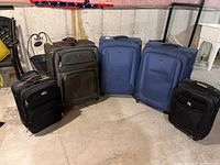 Five pieces of soft shell luggage, two blue and three black, all on wheels, arranged in a basement area showing size and condition.
