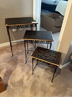 Photo showing all three nesting tables arranged by size, black tops with gold floral designs, metal legs, on carpeted floor.