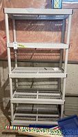 Front view of white plastic five-level shelf unit with grid shelves and cylindrical supports in basement setting.