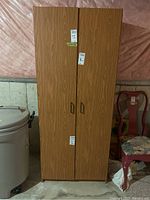 Front view of the MDF shelving cabinet with closed doors, showing wood grain finish and vertical handles, set in a basement environment next to a gray trash bin and a red wood chair.