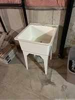 White plastic utility wash sink standing on concrete basement floor, adjacent to boxes and waste bin, view from angle showing basin and legs.