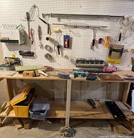 Wide angle view showing entire workbench with assorted tools, supplies, tapes, containers of fasteners, hand tools hanging on pegboard above bench, yellow plastic toolbox on bench, various hardware items scattered on and under workbench.