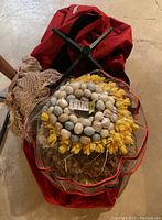 Red Christmas tree storage bag with zipper, black metal tree stand, and several wreaths stacked inside the bag, including one with yellow flowers and pastel colored eggs.