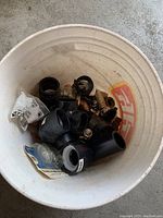 Top view of white bucket filled with black PVC pipe fittings, brass valves, and miscellaneous plumbing parts.