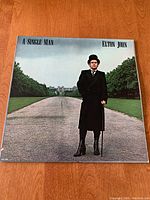 Front cover of vinyl album 'A Single Man' by Elton John showing artist in black coat with cane on pathway.