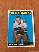 Front of 1965/66 Topps hockey card showing Glenn Hall in Chicago Black Hawks goalie uniform against bright blue background with Black Hawks team name.
