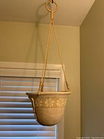 Front view of beige ceramic hanging pot with daisy flower decoration and natural fiber ropes, hanging indoors.