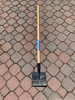 Full view of ice scraper on stone paver surface showing the entire wooden handle and black metal blade.
