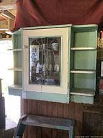 Front view of antique rustic wooden wall cabinet with painted green and cream finish. Central glass door with wood fretting and side open shelves. Cabinet mounted on wall with worn paint and aged wood visible.