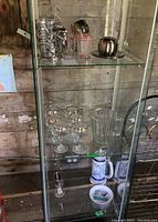 Photo showing six stem glasses with gold tone rims, clear ribbed glass pitcher, lower shelf with Avon beer stein, stem glass and novelty saucer, and metal figural beer mug on top shelf.