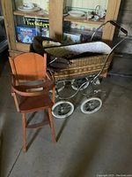 Full view of the wicker doll pram featuring a metal frame, white wheels, and a corduroy canopy next to the wooden doll high chair with tray and safety bar.