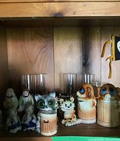Shelf with an arrangement of vintage salt and pepper shakers shaped like animals and human figures, plus transparent drinking glasses.