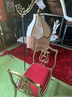 Photo shows tasseled floor lamp with ornate metal arm next to Baroque style sitting bench with red seat cushion and decorative wood backrest, highlighting wear on lamp shade.