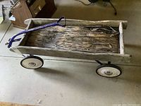 Full view of old wagon with metal frame, wooden bed, blue metal handle, and four wheels on a concrete floor.