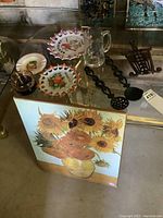 Wide view of all items together on glass table including souvenir plates, ashtrays, beer stein, iron spoons, and sunflower print plaque leaning against table.
