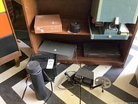 Full view of the vintage projectors, film editor, slide cases, and camera lenses arranged on a floor with a black and white patterned surface, and a wooden shelf in the background.