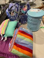 Overview photo of doll bedding items, two purple doll crutches, blue star patterned fabric bag with doll clothes, and turquoise mini dresser.