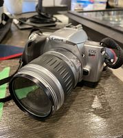 Front left angle view showing Canon EOS Rebel 77 SLR film camera body with attached zoom lens and camera strap on table surface.
