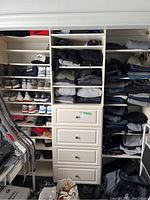 Full view of white closet shelving unit with shelves holding folded clothes and shoes, plus four drawers in center column.