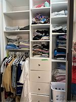 Full view of the closet shelving unit with clothes folded in the shelves, clothes hanging on the rod, and drawers visible in the middle.