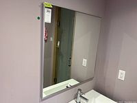 Front view of the square mirror mounted on pale purple wall above a sink, showing its clean reflective surface and metal frame.