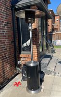 Full view of the outdoor propane patio heater standing on a paved patio area adjacent to a brick house wall. Shows overall structure and wheels at base.
