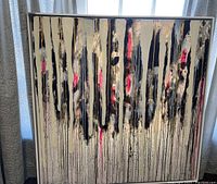 Frontal view of abstract painting with vertical drips in black, cream, pink, and gold tones, framed and standing upright near a window with curtains.