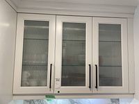 Wide front view of white upper cabinet with three vertical textured ribbed glass doors and black handles. Part of kitchen corner visible.