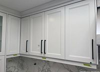 Front view of several white painted upper kitchen cabinet doors with black handles mounted in kitchen space. Shows clean paneled door style and black hardware.