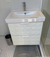 Front view of white wall mounted vanity with geometric drawers, ceramic sink, chrome faucet, and trash can beside it