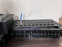 Front and top view of the black metal audio snake box featuring multiple XLR input and output ports and connectors, placed on a wooden surface near a wall.
