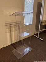 Clear acrylic podium with top surface, middle shelf, and solid base, placed in carpeted room near window with blinds.
