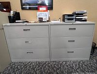 Front view of two grey metal file cabinets side by side, showing three drawers each with recessed handles. Some office items are placed on top of them.