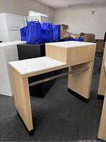 Photo angled showing front and side view of tall wooden desk with raised box compartment and lower desk surface, light wood with white tops and black trim.