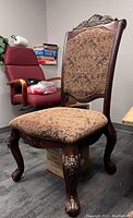 Photo of a wooden chair with a cushioned seat and backrest, ornate carvings on the wood frame, placed in an office room.