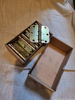 Cardboard box containing 18 used brass door hinges stacked inside, one hinge resting on top showing screw holes and hinge pin.