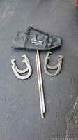Four metal horseshoes, two metal stakes, and a black fabric carrying bag on pavement.