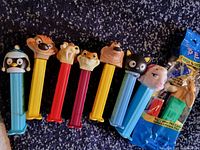 Photo shows eight Pez candy dispensers with character heads from animated movies. Seven dispensers are out of packaging, one is blue-packaged unopened. Characters include a penguin, Timon and Pumbaa from The Lion King, a cat, a pig, a hippo, and a lion.