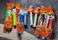 Eight PEZ dispensers arranged side by side on a gray surface, four are out of package and four are sealed in original orange packaging. Visible heads include ghosts, pumpkins, witch, and skeletons with glow in the dark themes.